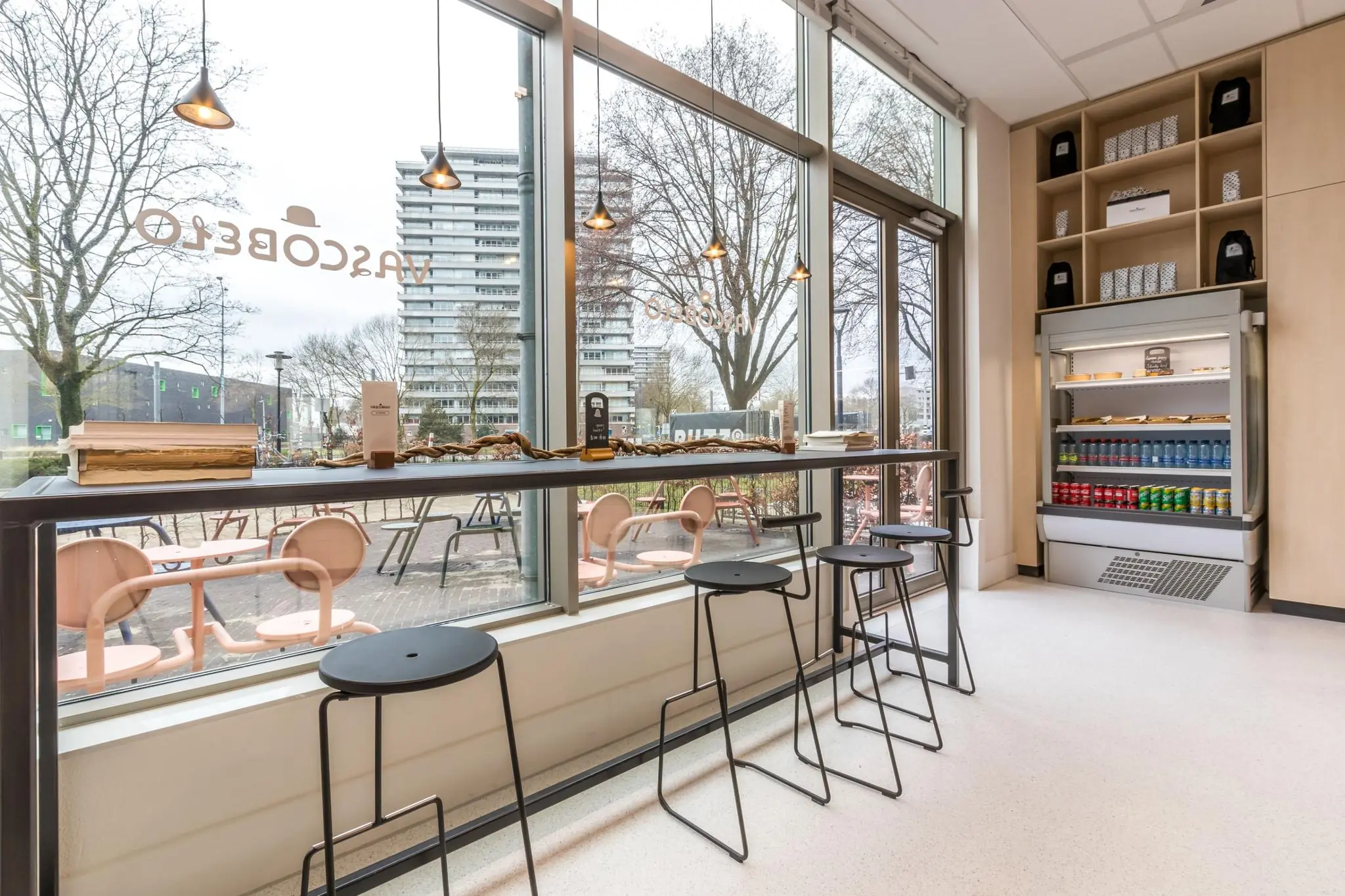 Modern interieur van een horecagelegenheid aan de Vliegend Hertlaan met hoge ramen, zitplekken aan het raam en een gekoelde displaykast met drankjes en snacks.